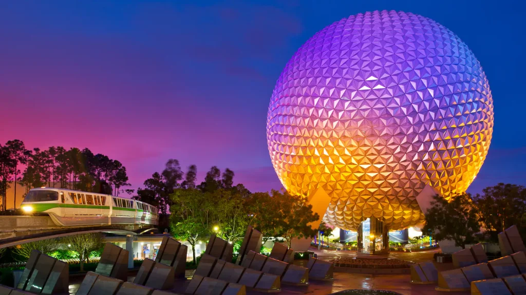 The EPCOT Ball illuminated at night in a purple glow with a monorail with a green stripe passing by. 