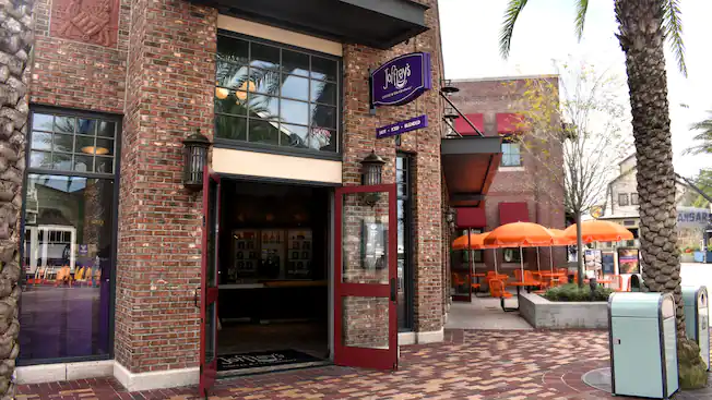 Outside of Joffrey's Coffee and Tea Company in Pixar Place in Hollywood Studios. Brick building with a blue sign. Red framed glass doors. Orange tables with orange umbrellas outside. 