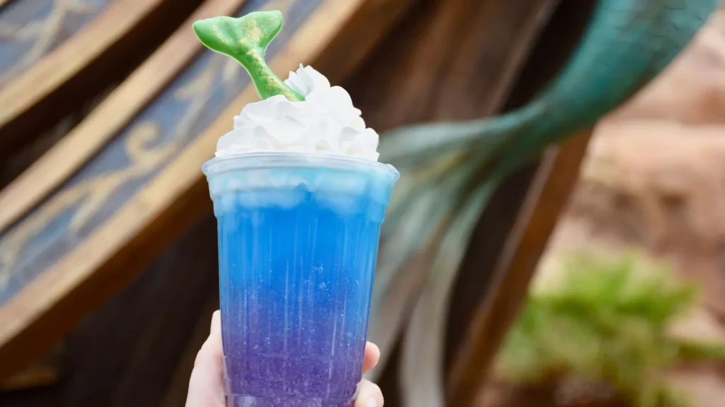 Blue and purple slushie with whipped cream and a Little Mermaid Tail poking out of the top. Available at Prince Eric's Village Market in Fanasyland.