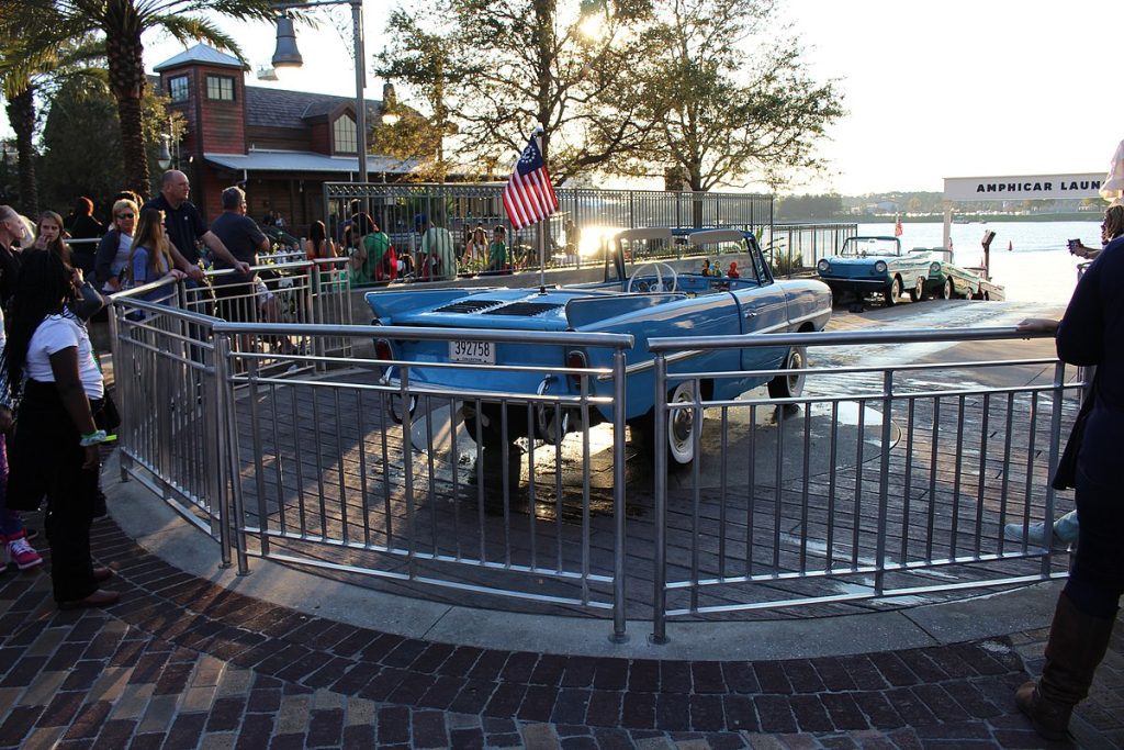 Amphicar Launch at Disney Springs. Blue, Convertible Amphicars Behind a Fence. Ramp for Amphicars to go in the water. Crowd of people watching.