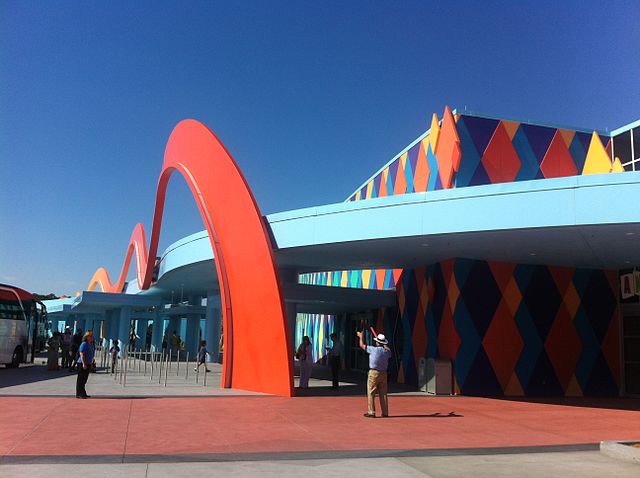 Art of Animation Hotel with Bus and People Outside. Queue fencing, multicolored designs on the walls of the hotel, and large orange arches.