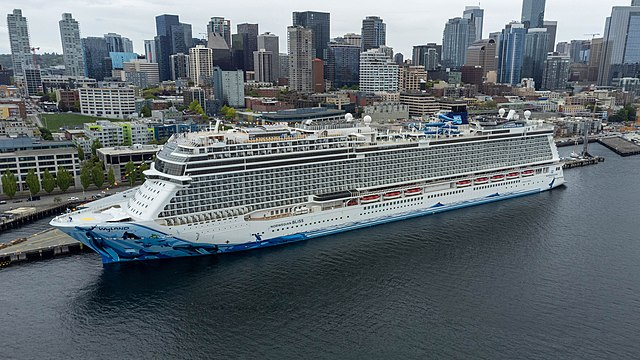 Norwegian Bliss Docked in Seattle Heading to Alaska. White ship, light blue waterslide, gray skyscrapers, dark blue ocean,